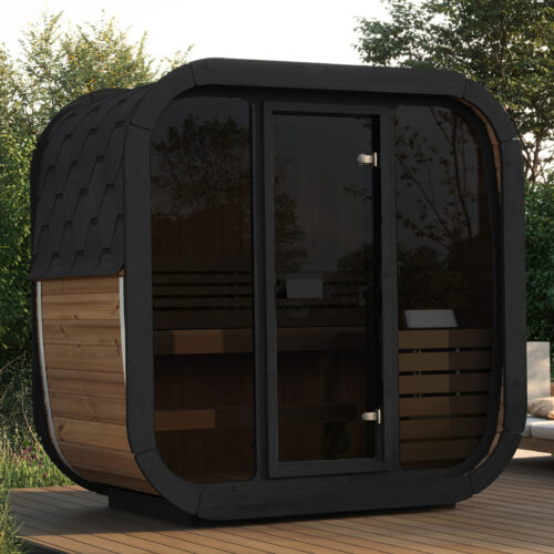 A small modern wooden sauna with tinted glass door on a wooden deck is surrounded by lush greenery. Nearby are cushioned chairs, creating a serene outdoor setting.